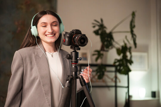 A young woman wearing headphones and a blazer smiles while recording with a camera on a tripod, creating content for her online audience.