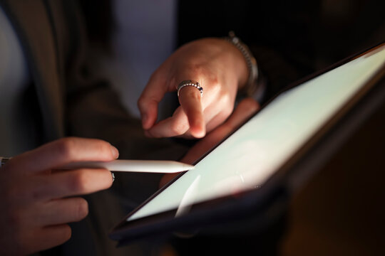 A person's hands are shown using a stylus to interact with a digital tablet. One hand holds the stylus, while the other touches the screen.