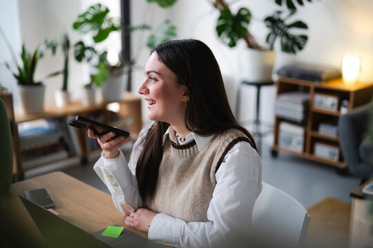 A young woman smiles while speaking on her phone in a bright, plant-filled home office.