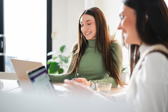 Two young women collaborate on a project using a laptop, sharing ideas and working together in a bright, modern office setting.