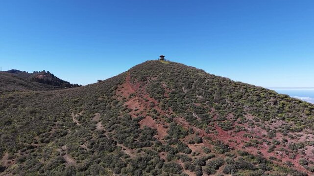 Drone flying around a remote fire watchtower on top of a mountain peak, offering a panoramic view of the surrounding landscape and ocean
