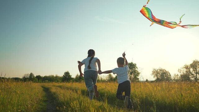 Girl and boy run holding hands flying rainbow kite in meadow at sunset outdoors. Sister with brother plays using colorful kite. Siblings run in field. Girl and boy fly kite. Children in meadow play.