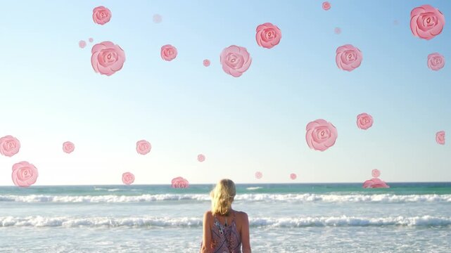 Multiple pink rose icons falling against rear view of caucasian woman standing on the beach