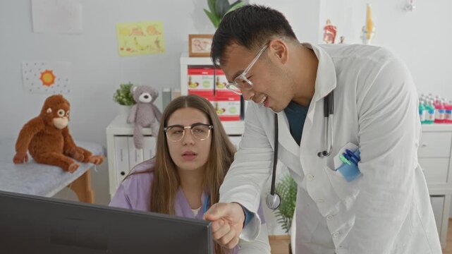 Man doctor in white coat points pen at computer screen while woman in scrubs watches and listens in pediatric exam building; teamwork reassurance.