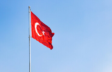 Turkish national flag with crescent and star waving on a silver flagpole against a clear blue sky background with green pine tree tops at the bottom in a bright daylight. Copy space. © Selim Aysolmaz