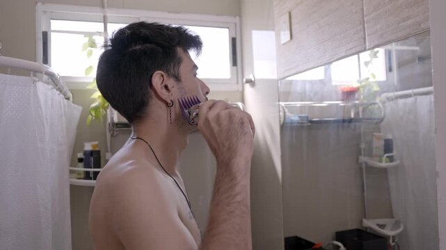 Shirtless young man with tattoos giving himself a haircut with an electric clipper in the bathroom, looking at his reflection in the mirror