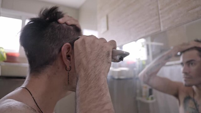 Young man with tattoos and earrings giving himself a haircut at home, using an electric razor in front of the bathroom mirror