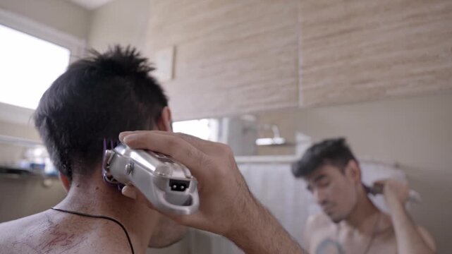 Young man giving himself a haircut at home using an electric clipper. He is looking in the mirror to carefully trim his hair