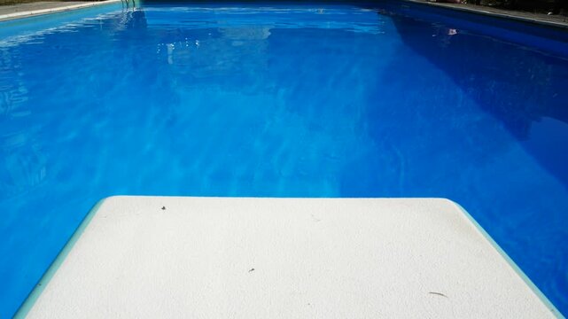 The diving board of a swimming pool in the foreground, with blue calm waters on the background. Outdoors view. 
