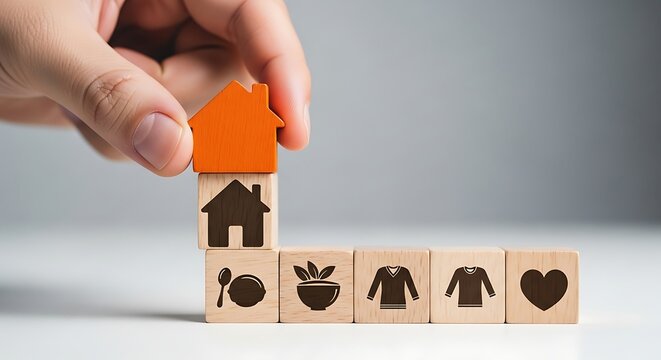 Hand stacking wooden blocks representing essential life needs and home ownership.