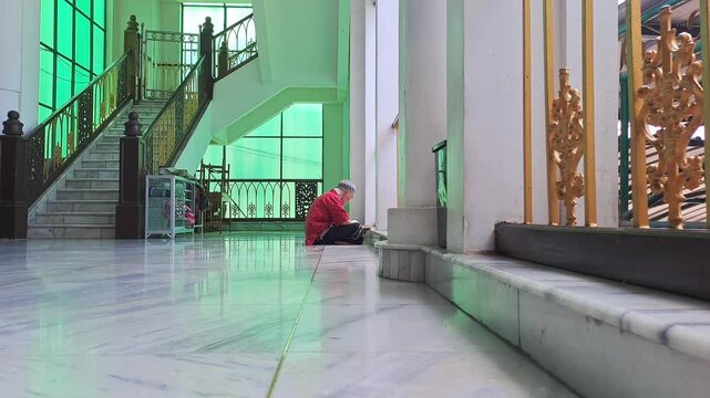 Close-up of a senior Asian Muslim man sitting in a corner of a mosque porch, opening and reading the Holy Quran. This serene footage captures the essence of aging with spiritual devotion.
