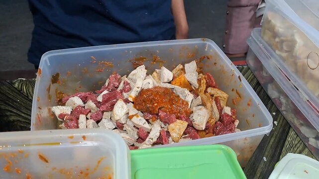 Close-up of an Indonesian street food vendor preparing Jigor, a popular snack made of fried sausages, batagor, and meatballs mixed with spicy chili sauce and lime leaves. The footage shows the vendor 