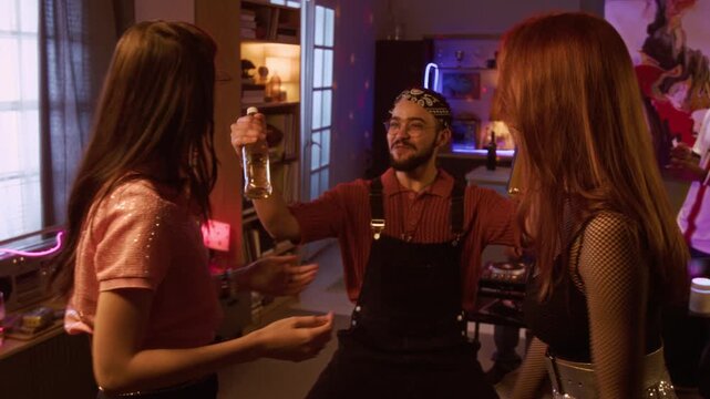 Pull back shot of two young adult women greeting with embrace during home party when cheerful biracial man bringing bottles with alcohol drinks, friends getting together for indoor celebration