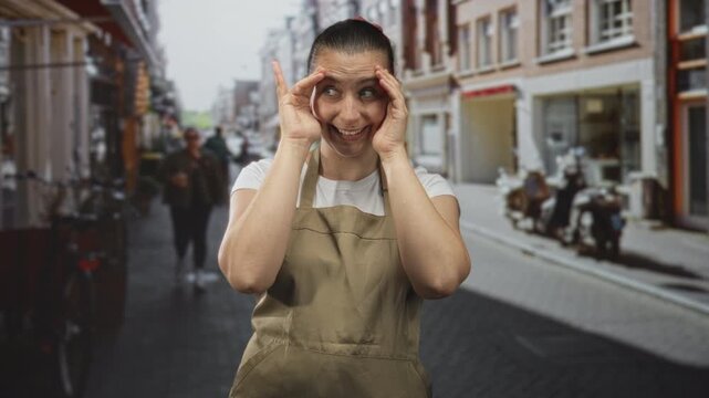 Woman in apron pressing hands to face and peeking through fingers on a busy street storefront with bicycle and motorbike visible; shyness.