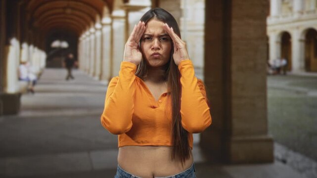 Woman covers face with hands and shows cropped midriff in arched building corridor wearing orange sweater; shyness curiosity.