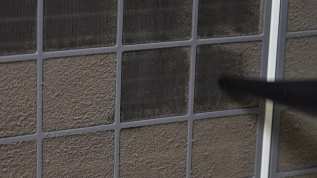 Black nozzle of a red handheld vacuum cleaner removing dust from a dirty air conditioner filter grid on a textured wall, home maintenance routine, appliance cleaning concept, domestic hygiene scene.
