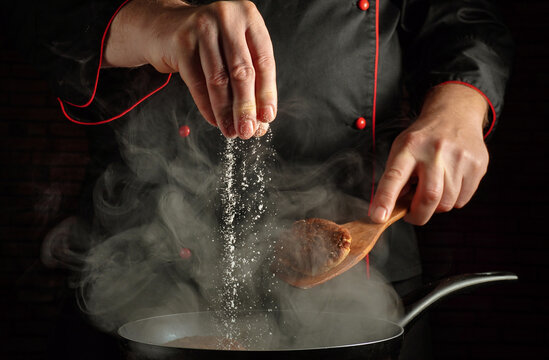 A chef sprinkles salt from a hand into a pot on the stove. Steam rises, showing the focus on cooking. The cook wears a dark coat with red trim
