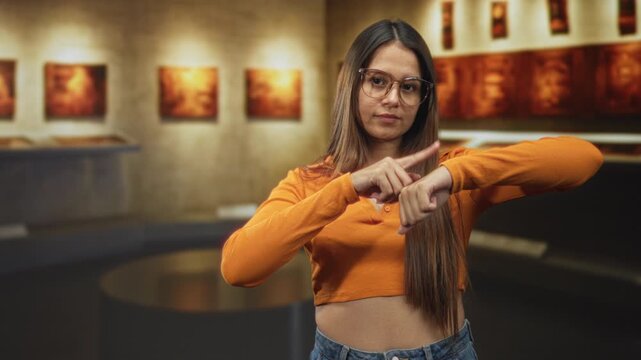 Woman points finger to wrist and shows midriff while wearing an orange crop top in a building gallery; impatience.