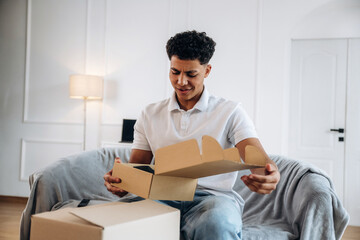 Fototapeta premium Young man with curly hair opening a cardboard box while sitting on a gray sofa in a modern living room with a lamp and desk in the background