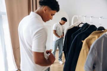 Fototapeta premium Young African American male in white polo shirt adjusts his outfit while standing in front of a mirror, clothing rack with various garments visible in the background