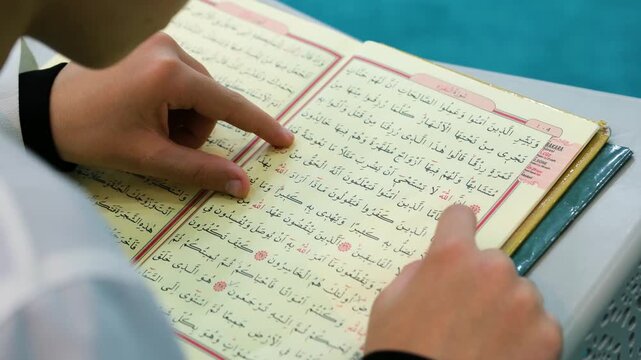 Teacher's hand guiding a student through the Arabic verses of the holy Quran for religious tajweed training and spiritual development at a mosque or islamic school during day