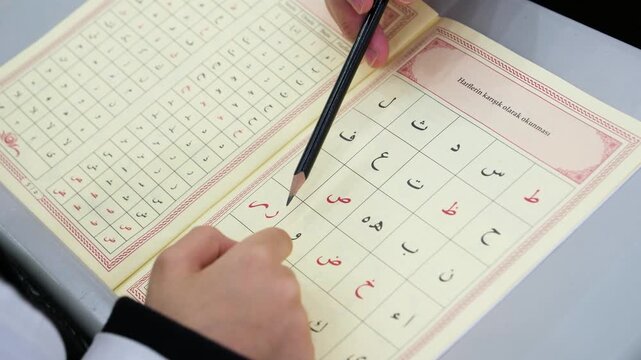 A teacher using a pencil to show specific Arabic letters on an Elifba page to a young student during a basic religious education lesson at an islamic school or mosque