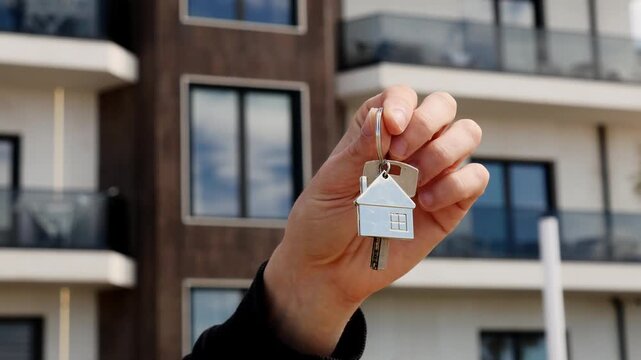 Key to new home with house-shaped keychain, symbol of property purchase and homeownership, new building visible behind.