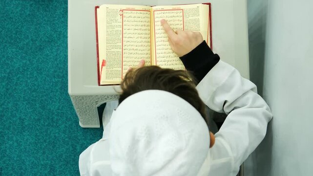Top view of a little boy sitting in a mosque and learning to read the Quran with focus and devotion for religious education and upbringing in the islamic faith during day