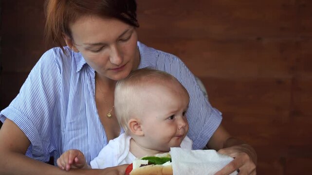 Hungry toddler sitting on mothers lap tries to take a bite of a large delicious sandwich at a restaurant.