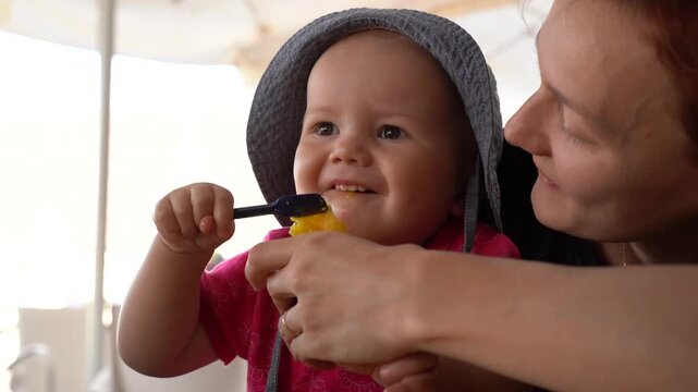 Happy baby girl sitting on mothers lap laughs and licks a spoon while eating delicious mango gelato at a cafe.