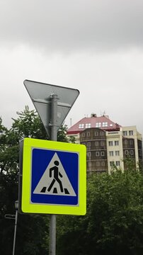 Fluorescent-bordered pedestrian crosswalk traffic sign beneath a triangular yield sign on a metal pole, set against leafy trees, overcast sky, and apartment buildings with red roof