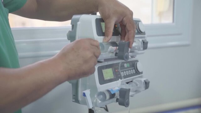 A side-profile, medium shot of a medical professional in green scrubs operating a dual-stacked syringe infusion pump. The professional's hands are shown precisely loading or adjusting a large syringe 