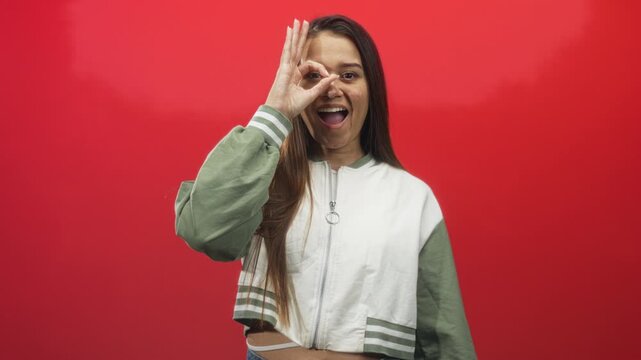 Woman makes ok hand circle over eye, smiling in a red studio wearing white and green zip jacket and jeans; playful confidence.