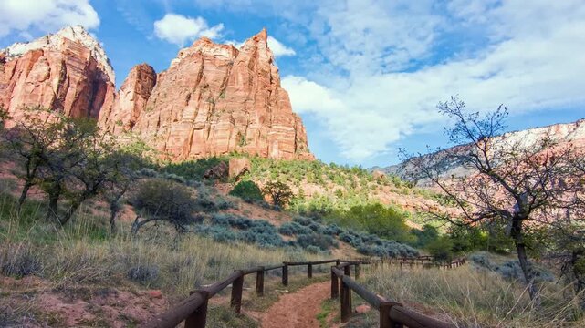 A beautiful view of the Sand Bench Trail in Zion National Park, Utah, featuring a dirt path with wooden fencing leading toward towering red rock cliffs and lush green vegetation under a blue sky with 