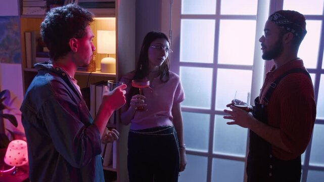 Pull back shot of young adult woman with cocktail glass in hand engaged in conversation with two male friends standing in living room during house party