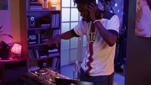 Tilt down shot of cheerful young adult Black man dancing to music while DJing during home party in neon lit apartment