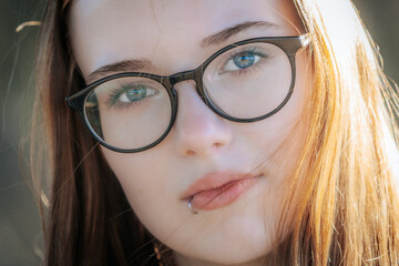 Fototapeta premium Young woman with long brown hair and glasses gazes directly at the camera, showcasing her blue eyes and subtle lip piercing against a softly blurred background