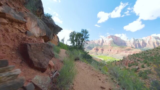 Footage captures the scenic Watchman Trail in Zion National Park, Utah, USA. The dirt path winds alongside a rustic stone wall with lush greenery, opening up to a breathtaking vista of massive red roc
