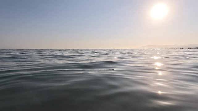 Cinematic low angle shot of gentle ocean waves under a bright sun. Calm sea surface with beautiful bokeh highlights, horizon line and soft golden hour light, creating a peaceful and serene seascape
