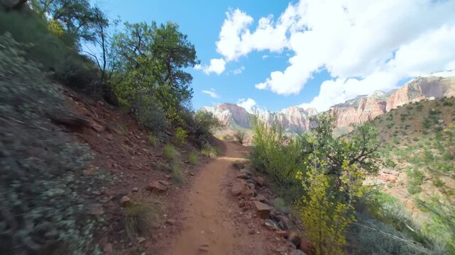 Footage of a hiker's point of view walking along the dirt path of the Watchman Trail in Zion National Park on a sunny day with blue skies and white clouds. The trail is surrounded by lush green vegeta