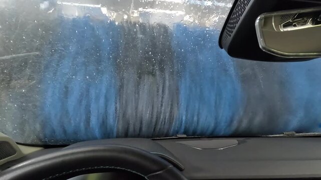 POV from inside a car during automatic wash tunnel, blue foam and water covering the windshield, dashboard visible, brushes and light outside