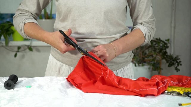 A person is skillfully cutting a piece of red fabric with scissors at a sewing station filled with tools. The setting is a bright, comfortable home space, showcasing creativity.