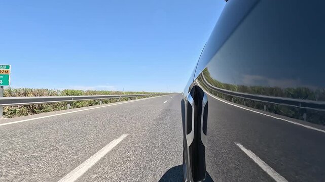Low angle view of a vehicle traveling at high speed on a two-lane road, with safety railings and clear blue sky, with reflections on the car