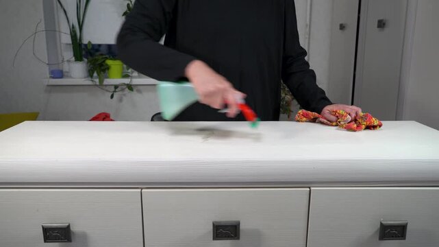 A person is actively cleaning a countertop using a spray bottle and cloth in a well-lit indoor environment. The space features various indoor plants and a minimalist design.