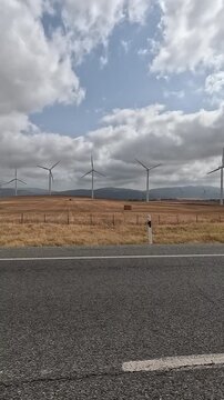 Vertical view of wind turbines on arid hills next to a rural road under a cloudy sky in Spain. Video taken from a vehicle traveling on a rural road