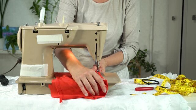 A person uses a vintage sewing machine to craft a vibrant red garment in a cozy workspace adorned with plants. Measuring tape and tools are nearby, creating a warm atmosphere.