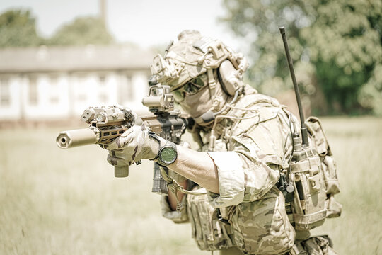 Soldier in tactical gear aims rifle with suppressor and optics in grassy field