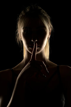 Mysterious female silhouette in black lingerie making a &ldquo;shh&rdquo; gesture with finger on lips, dramatic rim lighting on dark background