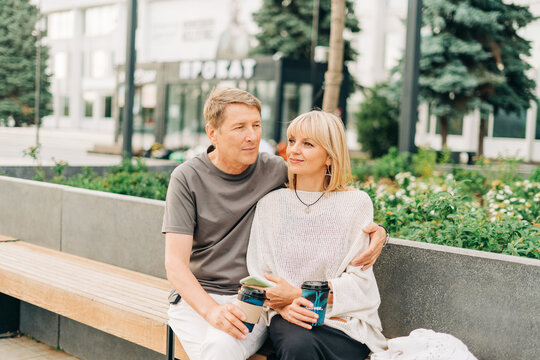 An adult mature happy couple in love sitting on bench outdoors in city street park. A blonde caucasian man and woman spend time together and drinking coffee. Senior wife and husband walking outside