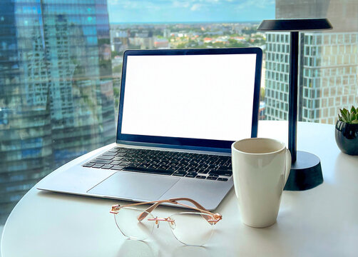 Modern workspace with laptop featuring blank white screen mockup, coffee cup and eyeglasses on table near window with city view. Clean minimalist composition with copy space, ideal for business, remot